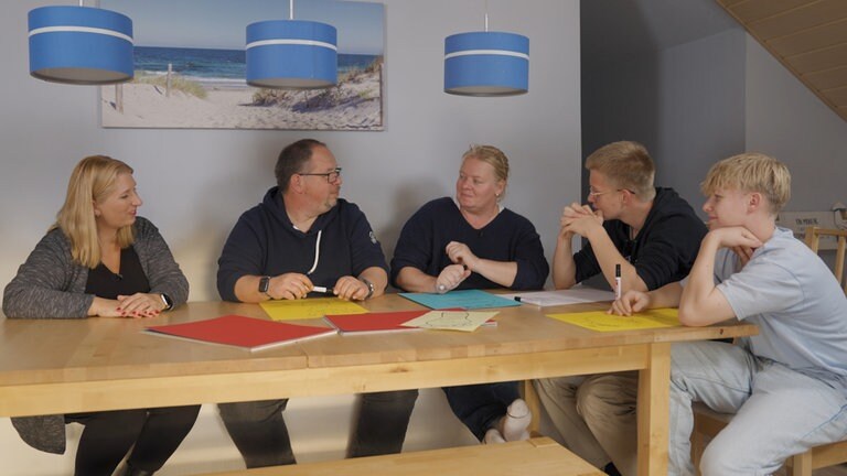 Fünf Personen sitzen an einem Esstisch, über dem Tisch hängen drei blaue Lampen. An der Wand hängt ein Bild von einem Strand. 
