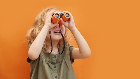 Ein Mädchen steht vor einer orangenen Wand und blickt mit einem orangenen Fernglas nach oben.