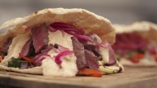 Ein Fladenbrot, in dem rosane Steakstücke, eine weiße Soße und rote Zwiebeln sowie Grillgemüse drin sind.