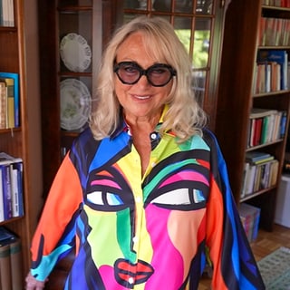 Sylvia ist 77 Jahre alt. Sie steht in ihrem Wohnzimmer und trägt eine auffällige, bunte Bluse mit einem Gesicht als Motiv. Außerdem trägt sie eine große, auffällige Sonnenbrille. 
