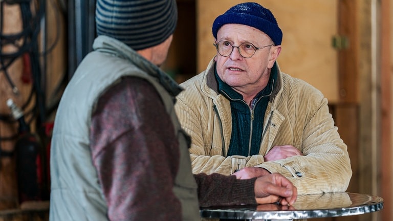 Toni (Roland Frey) lehnt an einem Stehtisch und unterhält sich mit Niki (Nikolaus König)