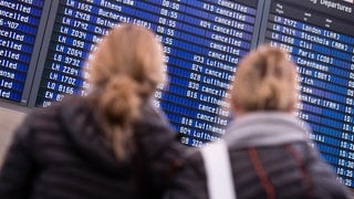 Zwei langhaarige Personen stehen mit dem Rücken zur Kamera zu einer Fluganzeigetafel blickend. Ein Großteil der Flüge ist dabei annulliert.