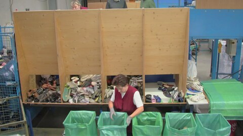 In einer Altkleider-Sortieranlage sieht man vier Schächte durch die Schuhe und Sneaker in verschiedene Kategorien sortiert werden. Eine Frau sortiert die Schuhe in Plastiksäcke.