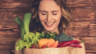 Junge Frau blickt lächelnd in braune Papiertüte, die bis zum Rand mit Gemüse gefüllt ist. Anti Aging: Welche Lebensmittel sind gut gegen Hautalterung?