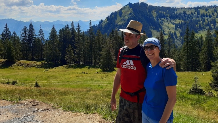Ein Mann und eine Frau vor einer grünen Wiese, im Hintergrund ein Berg und blauer Himmel.