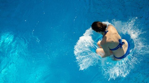 Die Saison zum draußen Schwimmen geht wieder los. Eine junge Frau spingt in das Wasser eines Freibadbeckens.