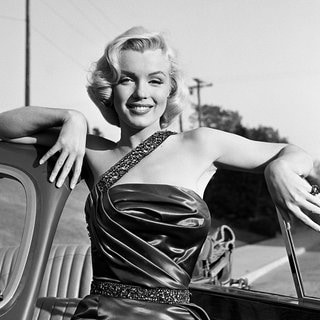 Marilyn Monroe lehnt sich lächelnd an ein Sportauto.