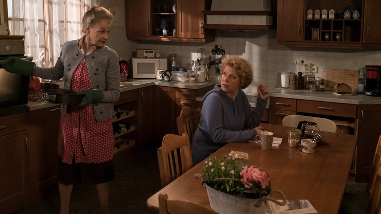 Bea sitzt am Küchentisch, Johanna holt einen Kuchen aus dem Ofen