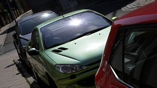 Ein dunkles, ein grünes und ein rotes Auto parken am Straßenrand. Die Sonne scheint auf die Windschutzscheiben und wird gespiegelt. 