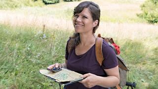 Eva auf einer blühenden Wiese, in der Hand eine Wanderkarte