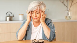 Frau sitzt am Tisch und reibt sich die Schläfen: Wenn es plötzlich warm oder kalt wird, können z.B. Migräne- oder Bluthochdruck-Patienten darunter leiden. Wetterwechsel können unserem Körper zu schaffen machen, weil er sich zunächst anpassen muss. 