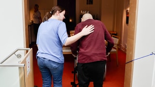 Melanie (links) arbeitet als Pflegerin im Hospiz. Sie läuft mit einem Gast (rechts) zum Esstisch, die beiden sind von hinten zu sehen. Melanies Arm liegt auf dem Rücken des Mannes.