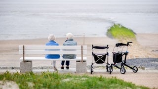 Zwei ältere Frauen sitzen bei sonnigem Wetter auf einer Bank an der Strandpromenade am Weststrand der Insel. Neben der Bank steht ein Rollator. Im Hintergrund sieht am den Strand und das Meer.