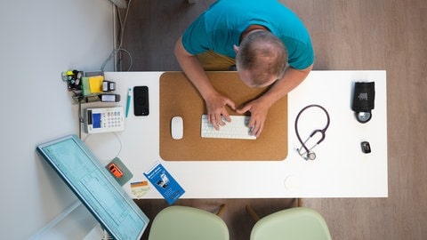 Ein Arzt sitzt an seinem Schreibtisch und tippt etwas auf seiner Tastatur ein. Eingeführt wurde sie bereits Ende April. Nun wird sie zur Pflicht: Ärztinnen und Ärzte müssen ab Oktober Diagnosen und Befunde in die elektronische Patientenakte (ePA) eintragen.