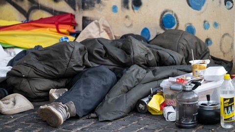 Ein Schlafsack bedeckt eine obdachlose Person. Daneben stehen Essen und Trinken. Für Obdachlose gibt es in Tübingen mehrere Unterkünfte.
