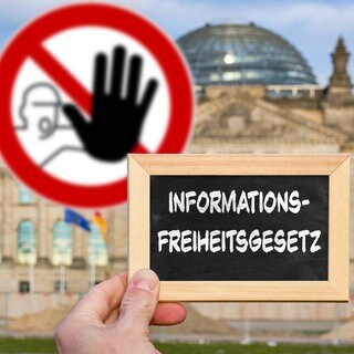Hand mit Tafel vor dem Reichstagsgebäude, darauf der Schriftzug "Informationsfreiheitsgesetz". Im Hintergrund ein Warnschild mit erhobener Hand: Um die Informationsfreiheit steht es in Deutschland nicht besonders gut