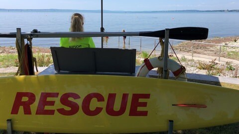Eine Frau sitzt unter einem Sonnenschirm und blickt auf einen See.