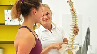 Physiotherapeutin erklärt einer Patientin die Rückenproblematik mithilfe eines Wirbelsäulenmodells: Die Kräftigung der Muskeln erfordert spezielle Krankengymnastik. Die Schultergürtelmuskulatur hat die Eigenschaft hat, in bestimmten Regionen zu verkürzen. So kommt es etwa durch Schreibtischtätigkeit zur Muskelverkürzung und in diesen Bereichen verkürzter Muskeln dann gehäuft zu Blockierungen. Daher ist es notwendig, die verkürzten Muskelgruppen zu dehnen.