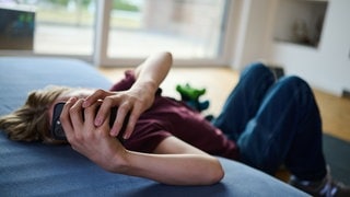 Ein Teenager guckt auf ein Handy, während er auf Boden und Matratze liegt. Das Handy verdeckt das Gesicht.
