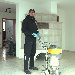 Andreas Röhrich steht in Arbeitskleidung in einem leeren Raum mit einem großen Gerät. Damit kann man schwere Verschmutzungen entfernen. 