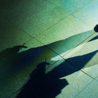 Der Tod - Im Anschnitt sind die Beine eines Mannes zu sehen; seine Erscheinung ist durch lange, ins Bild ragende und sich überlappende Schatten zu erkennen.
