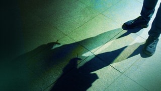 Der Tod - Im Anschnitt sind die Beine eines Mannes zu sehen; seine Erscheinung ist durch lange, ins Bild ragende und sich überlappende Schatten zu erkennen.