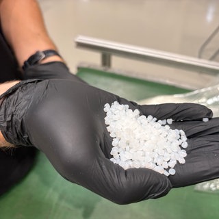 Weiße Kügelchen in einer Hand mit schwarzem Plastikhandschuh: Aus diesem Material entsteht das Glas, das weltweit gefragt ist. Die Freiburger Firma Glassomer hat ein spezielles Verfahren zur Glasherstellung erfunden. 