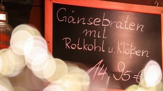 Ein Schild weist auf Gänsebraten hin (Symbolbild). Gans oder Fisch? Kurz vor Weihnachten boomen Festtagsessen. Doch Vogelgrippe und Personalmangel stellen Erzeuger vor große Herausforderungen.