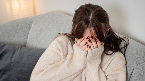 Eine Frau sitzt auf einer Couch und hält sich die Hände vor ihr Gesicht.
