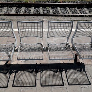 Eine leere Bank steht in der Sonne am Bahnsteig und wirft Schatten. 