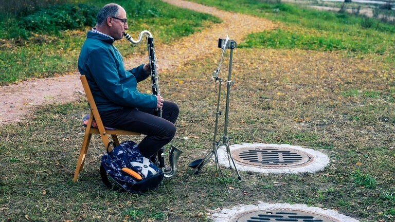 Mann auf Klappstuhl spielt Bassklarinette im Freien vor einem Gully