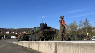Ein Soldat in Uniform, vermummt und mit Warnweste steht vor einem Panzer. Er wartet, bis der Güterwagon frei wird.