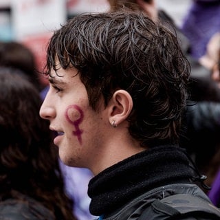 Junger Mann bei einer Demonstration, auf der Wange trägt er das Gender-Symbol für Weiblichkeit (Venussymbol): Können Männer Feministen sein?