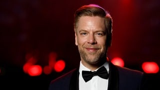Der Sänger Tom Gaebel steht in einem schwarzen Anzug und mit schwarzer Fliege vor einem rot-schwarzen Hintergrund und lächelt in die Kamera.