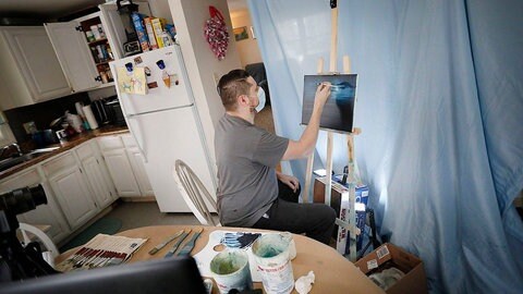 Ein Mann sitzt mit Maske vor einer Leinwand und malt ein Bild.