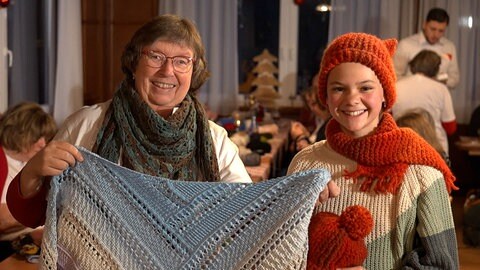 Eine Frau der flotten Nadeln zeigt gemeinsam mit einer jungen Interessentin ihre Strickartikel. Darunter zwei Schals und eine Mütze. 