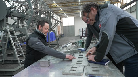 Drei Männer lehnen über einem Arbeitstisch in der Fabrikhalle des Gundelfinger Fassadenherstellers Josef Gartner GmbH. Sie arbeiten am Glaskreuz der Kathedrale Sagrada Família in Barcelona. Im Sommer 2026 soll das Bauwerk fertig sein.
