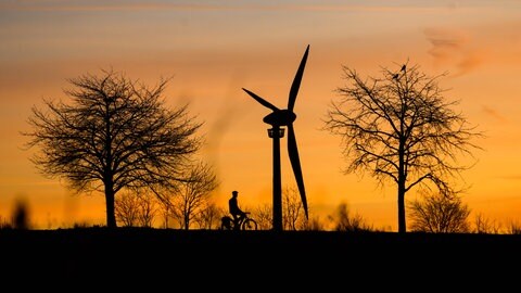 Ein Mann fährt bei Sonnenaufgang mit einem Fahrrad vor einem Windrad vorbei: 2024 stagnierte der Ausbau der Windkraft in der Südpfalz und Vorderpfalz
