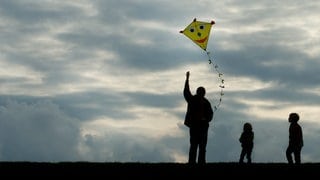 Ein Vater lässt mit zwei Kindern einen gelben, lachenden Drachen in den Himmel steigen - die Herbstferien in RLP starten.