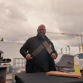 Grillmeister Ralf Ahrens steht am Grill und hält zwei Pfannenwender in der Hand.