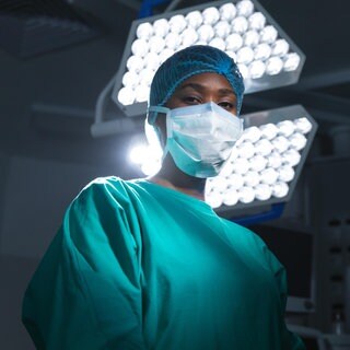 Schwarze Chirurgin mit Maske und OP-Kittel steht unter einer OP-Lampe: Krankenhäuser und Pflege-Einrichtungen sind auf medizinisches Personal aus dem Ausland angewiesen. 