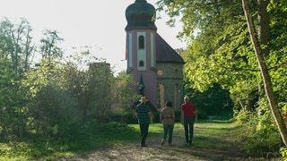 Drei Menschen gehen auf die Kirchenruine Mariahilf in Mühlheim zu. Sie steht mitten im Wald. 