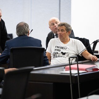 Der Querdenken-Initiator Michael Ballweg im weißen T-Shirt sitzt mit Rechtsanwälten in einem Saal des Landgerichts Stuttgart. Ballweg steht wegen versuchten Betrugs vor Gericht. (Archivbild)