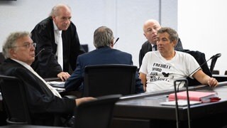 Der Querdenken-Initiator Michael Ballweg im weißen T-Shirt sitzt mit Rechtsanwälten in einem Saal des Landgerichts Stuttgart. Ballweg steht wegen versuchten Betrugs vor Gericht. (Archivbild)