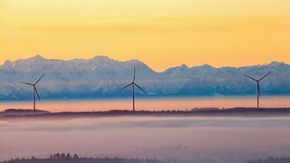 Drei Windräder ragen am Morgen kurz nach Sonnenaufgang aus der Nebeldecke, während im Hintergrund durch den Föhn die über einhundert Kilometer entfernten Alpen zu sehen sind.