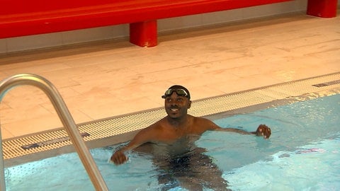 Ein Mann sitzt entspannt im Wasser am Beckenrand und lacht in die Kamera. Vor drei Jahren wäre Romeo Ayemele Djeujo fast ertrunken. Jetzt trainiert er selbst für das Rettungsschwimmabzeichen Gold.