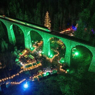 Ein Weihnachtsmarkt im Schwarzwald findet unter einem grün beleuchteten Viadukt statt. 