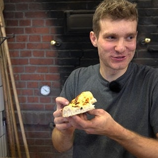 Ein junger Mann steht vor einem Backsteinofen und hält einen Zwiebelkuchen in der Hand. Mit seiner Leidenschaft für Pizza hat sich der Student Jonathan Brodbeck mit einem Catering selbstständig gemacht.