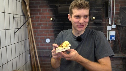 Ein junger Mann steht vor einem Backsteinofen und hält einen Zwiebelkuchen in der Hand. Mit seiner Leidenschaft für Pizza hat sich der Student Jonathan Brodbeck mit einem Catering selbstständig gemacht.