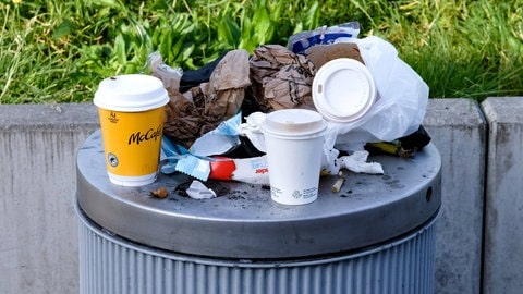 Auf einem vollen Mülleimer stehen leere Einweg-Kaffeebcher (Symbolbild).